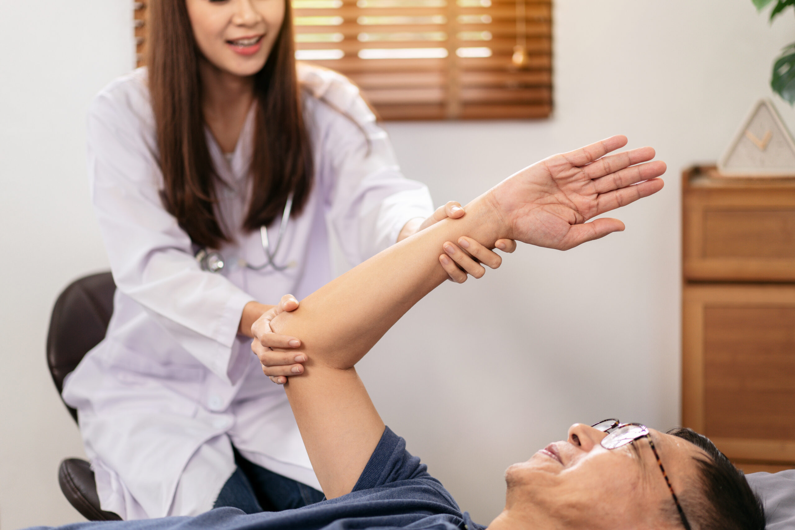 Female physical therapist working and doing arm rehabilitation therapy for elderly patient lying on hospital bed and talking to giving advice on treatment to patient.
