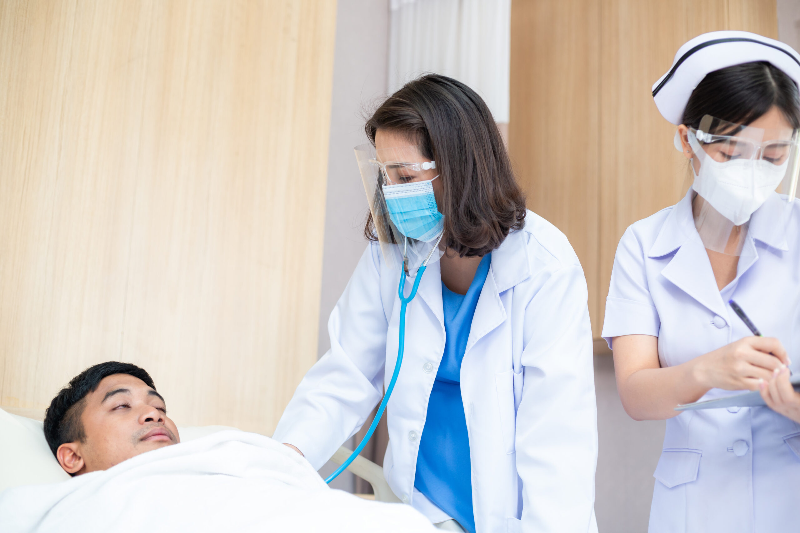 Asian female doctor wearing mask and face shield with nurse using stethoscope checking health of patient sleeping in bed at hospital ward