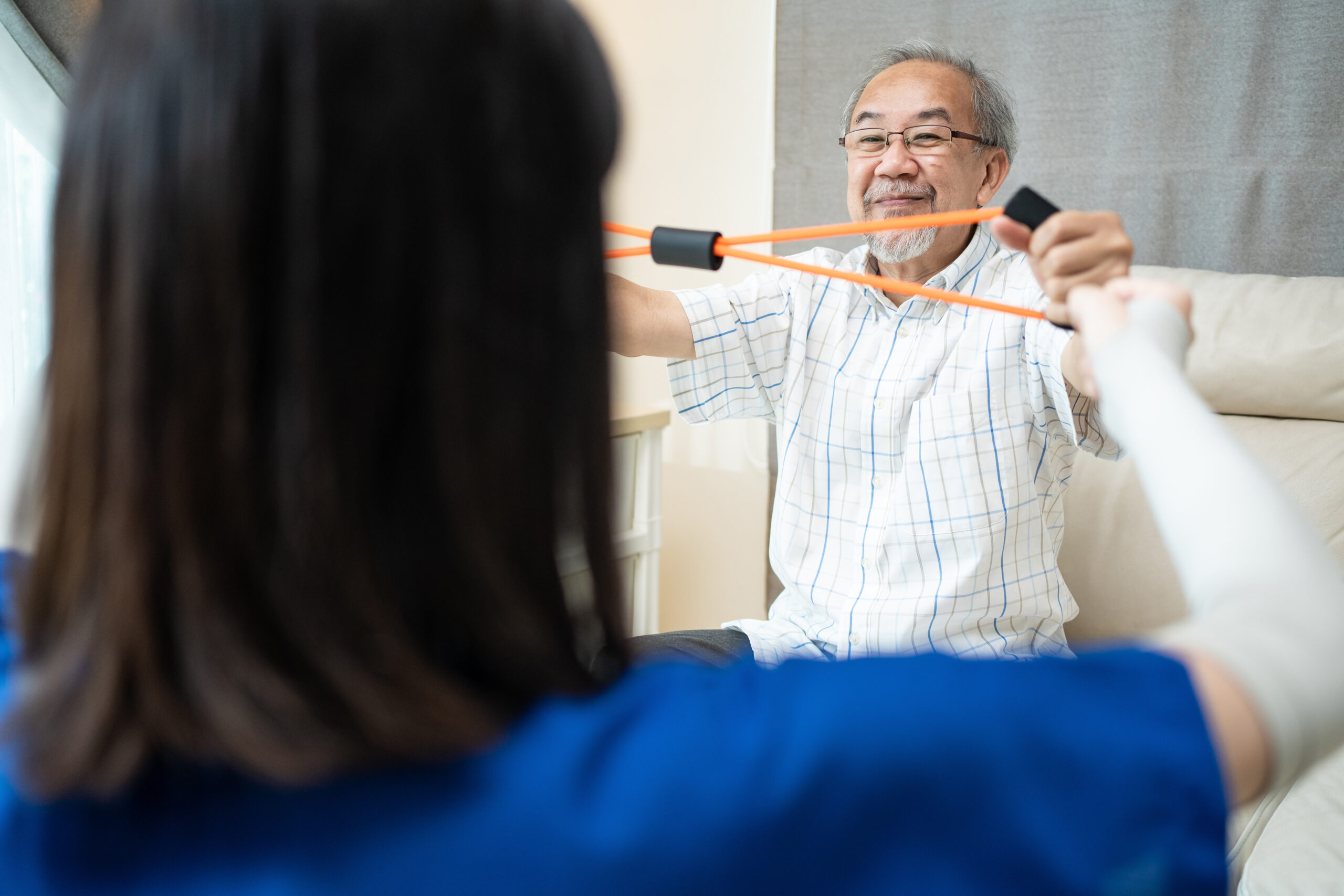 Asian Disabled senior older man sitting on sofa doing physiotherapist. Elderly handicapped man using resistance stretch band exercise for patient with support from therapist nurse in home nursing care